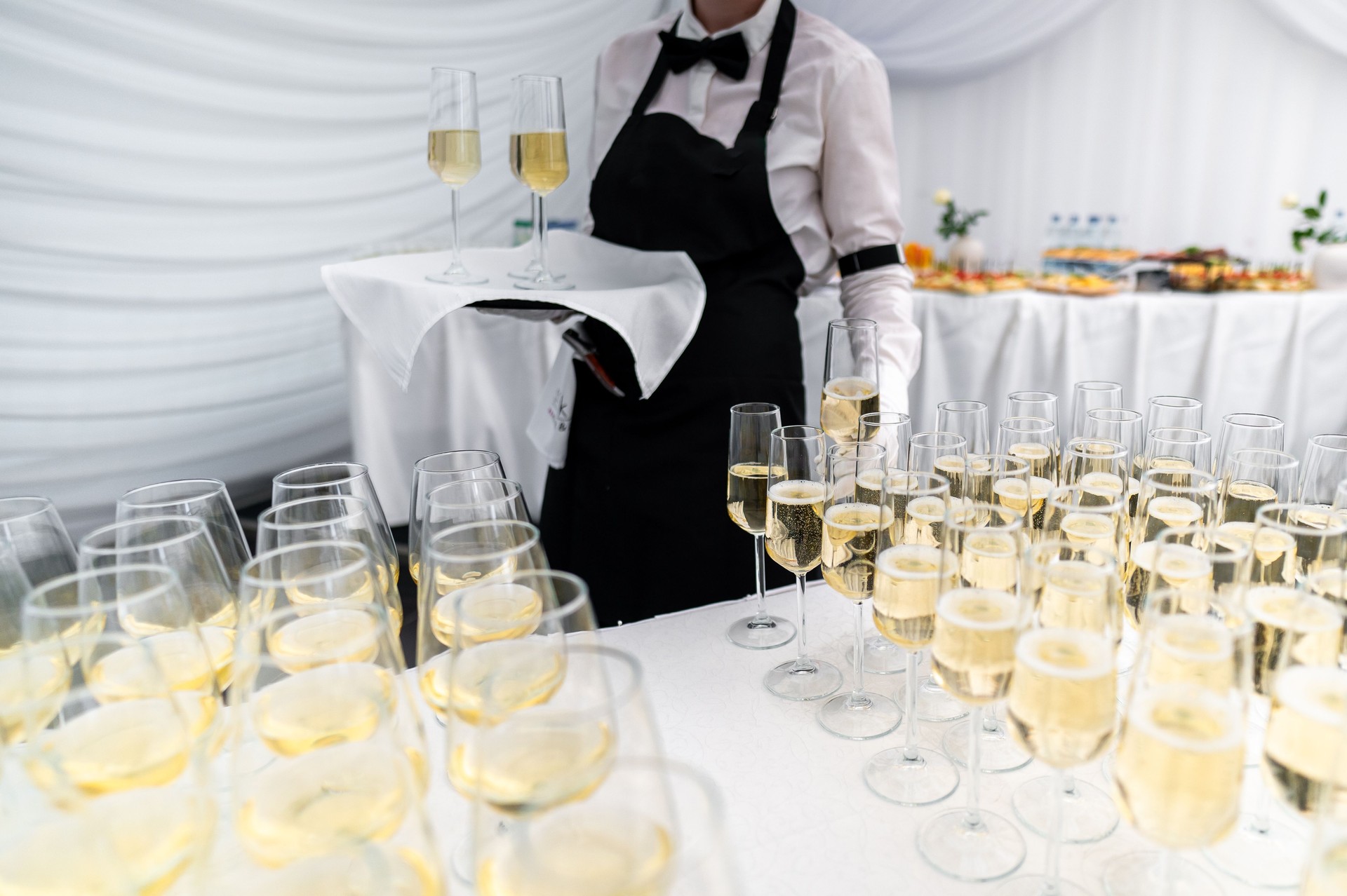 A waiter in a black vest and white shirt holds a tray of champagne glasses at an elegant event. In background, a table is set with various appetizers, creating a sophisticated and inviting atmosphere.